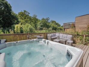 A hot tub with a wicker sofa and table, surrounded by a wooden deck and lush greenery.