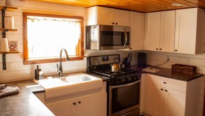 A compact kitchen with white cabinets, a stainless steel microwave, a gas stove, and a sink. - Charming Cabin + Pond View + Hot Tub (Carthage)