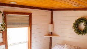 A bedroom with a quilted bedspread, a bedside lamp, a window with blinds, and a wreath on the wall. - Charming Cabin + Pond View + Hot Tub (Carthage)