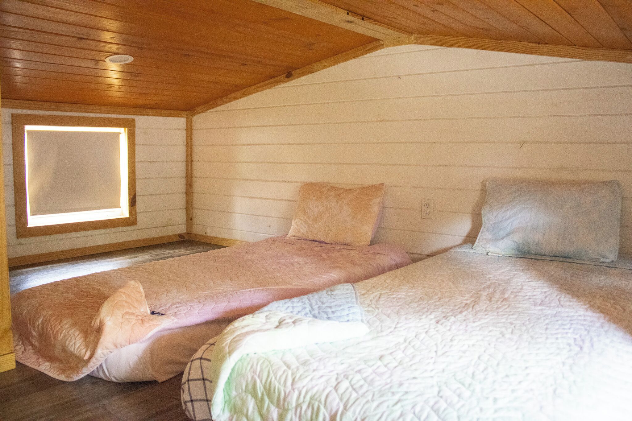 A small, cozy room with wooden walls and a sloped ceiling. There are two beds with pink and white bedding. A window provides natural light.