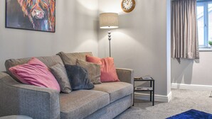Un salon avec un canapé gris, une lampe, une horloge et une fenêtre avec des rideaux.