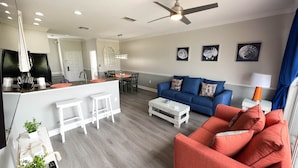 A modern living room with a blue sofa, red armchairs, and a white coffee table. There is a kitchen area with a dining table and chairs.
