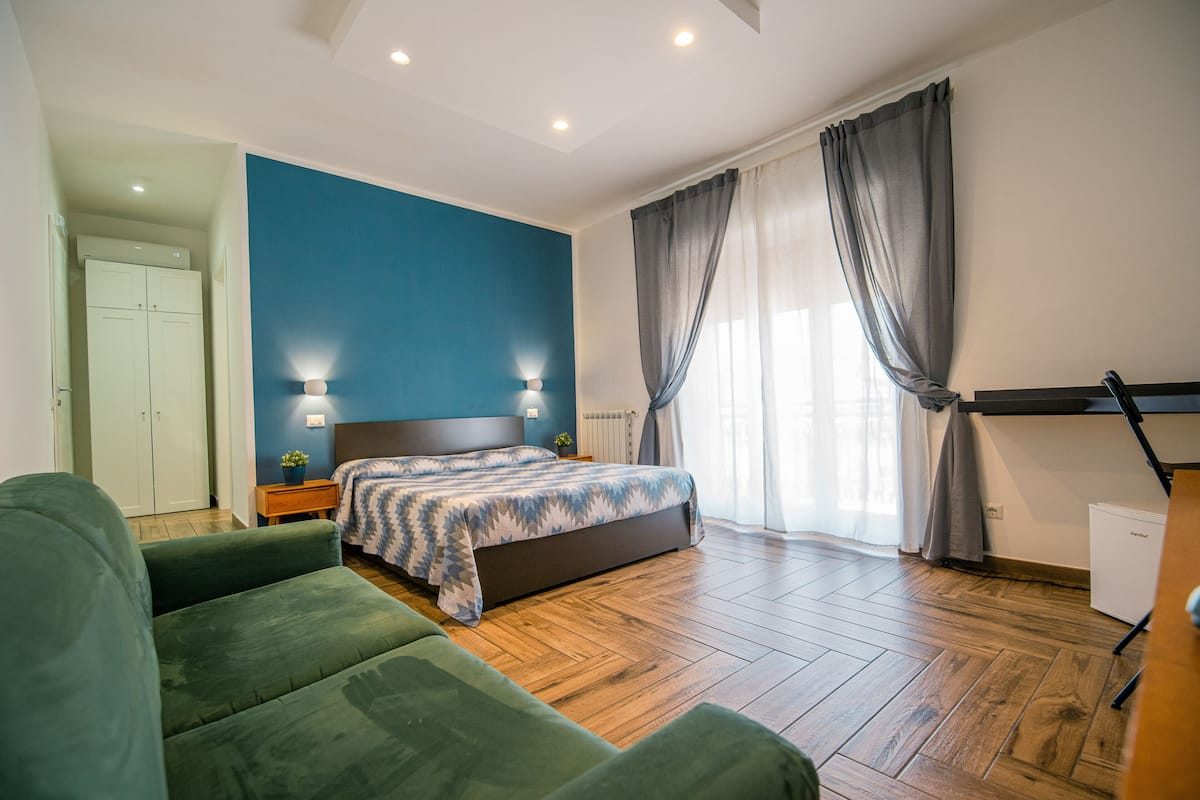 Un dormitorio con cama, sofá verde, suelo de madera y una ventana con cortinas.