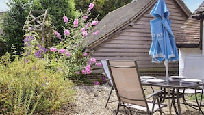 A wooden building with a sloped roof, a garden with gravel paths, and a table with chairs and a blue umbrella.