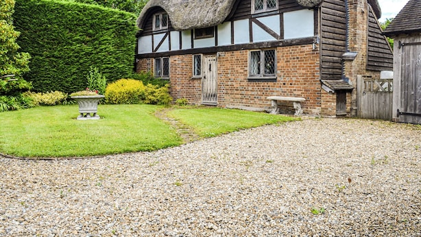 A traditional half-timbered house with a thatched roof, a gravel driveway, and a well-maintained garden with a stone bench and a flower bed.