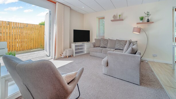 A modern living room with a large flat-screen TV, grey sofas, a coffee table, and a wooden deck visible through the sliding glass door. - 1 Ladyknowe - UK39970 (Moffat)