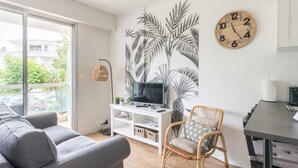 Un salon moderne avec un canapé gris, un fauteuil en osier, un téléviseur posé sur un meuble blanc, une horloge murale et une grande fenêtre donnant sur des immeubles et des arbres.