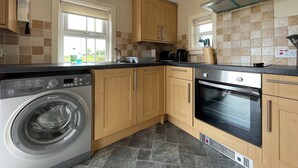 A compact kitchen with wooden cabinets, a built-in washing machine, a stove, and a window with a view. - Birch Cottage - UK38992 (Dalcross)
