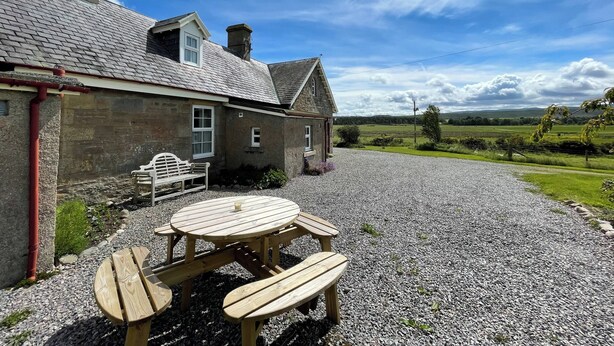 A stone cottage with a gravel driveway and a wooden table and bench set outside. - Birch Cottage - UK38992 (Dalcross)