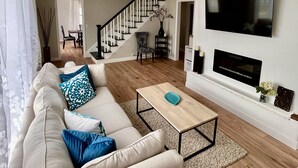 Een woonkamer met een bank, salontafel, televisie, open haard en een trap.