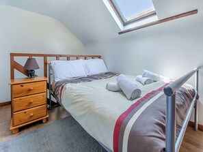 Un dormitorio con tragaluz, una mesita de noche de madera con lámpara, una cama con toallas enrolladas y una alfombra gris.