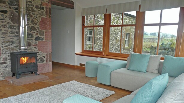 A cozy living room with a stone wall, a wood-burning stove, a sofa with teal cushions, and a view of a stone building through large windows.