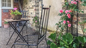 A rustic outdoor seating area with a metal chair and a wooden table, surrounded by potted plants and blooming flowers against a brick wall.