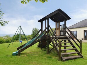 A playground with a slide and swing set, wooden climbing structure, and a house in the background. - 3 bedroom accommodation in Dunterton, near Tavistock (Dunterton, near Tavistock)