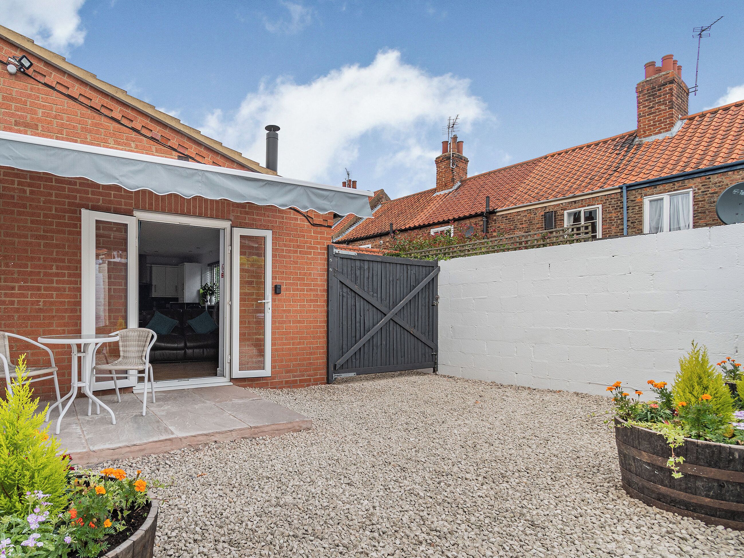 A patio area with a gravel ground, a white fence, and a brick building with a tiled roof.
