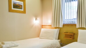 Een hotelkamer met twee enkele bedden, een houten hoofdeinde, een nachtkastje, een lijst met een afbeelding aan de muur en een raam met gordijnen.