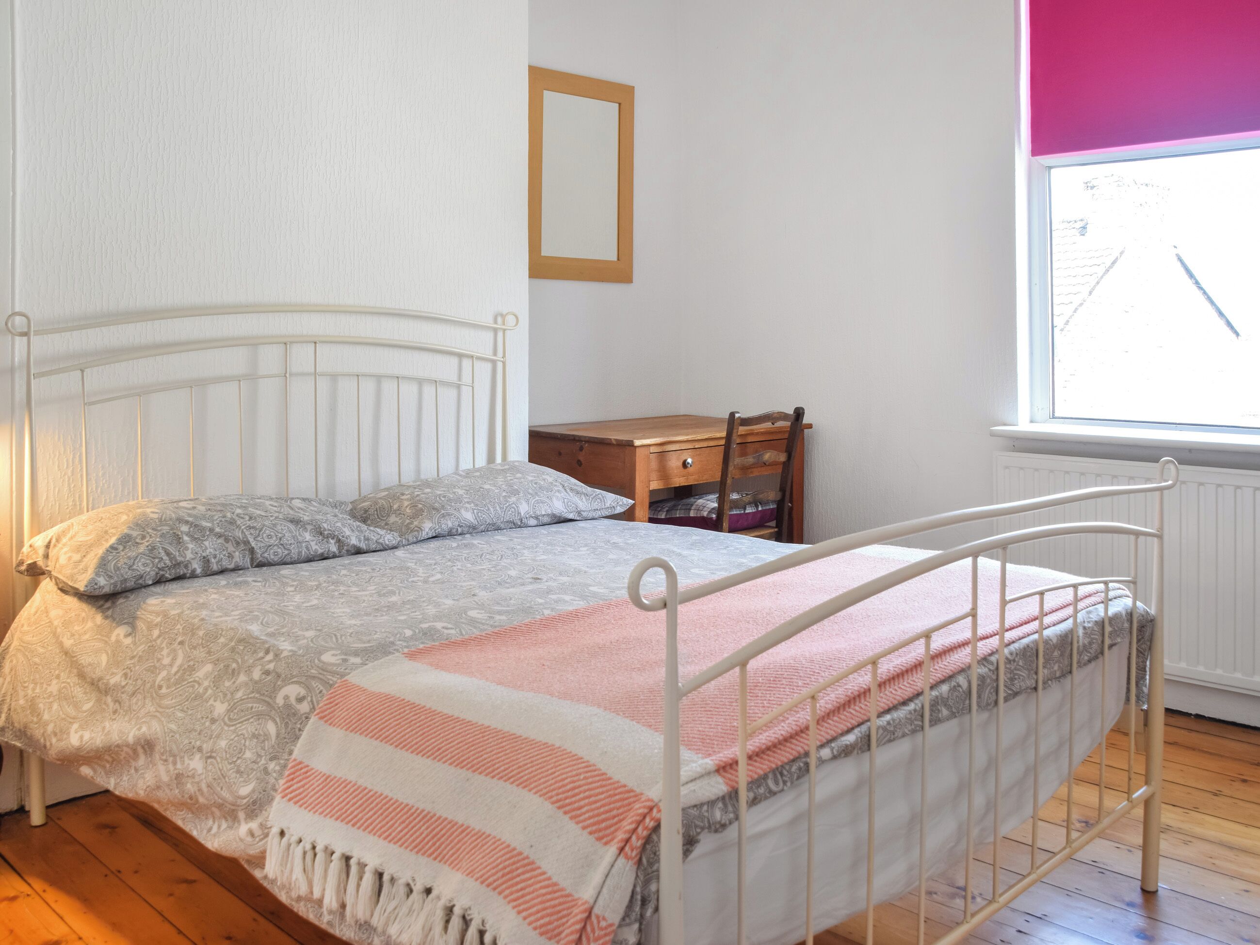 Un dormitorio con cama, mesita de noche de madera, una silla, ventana y radiador.