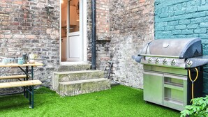 Un bâtiment en briques avec une pelouse artificielle verte, un barbecue et une table en bois sur laquelle se trouvent des tasses et une cafetière.