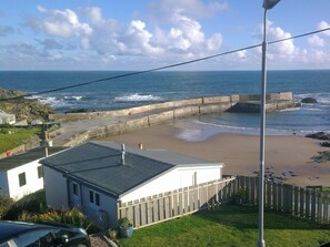 A coastal view with a sandy beach, a wooden fence, and a building with a grey roof.