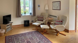 A cozy living room with a television, a fireplace, two white armchairs, a patterned rug, and a window with curtains. - Detached cottage on the banks of Loch Long and the Firth of Clyde near Dunoon. (Ardentinny, Dunoon)