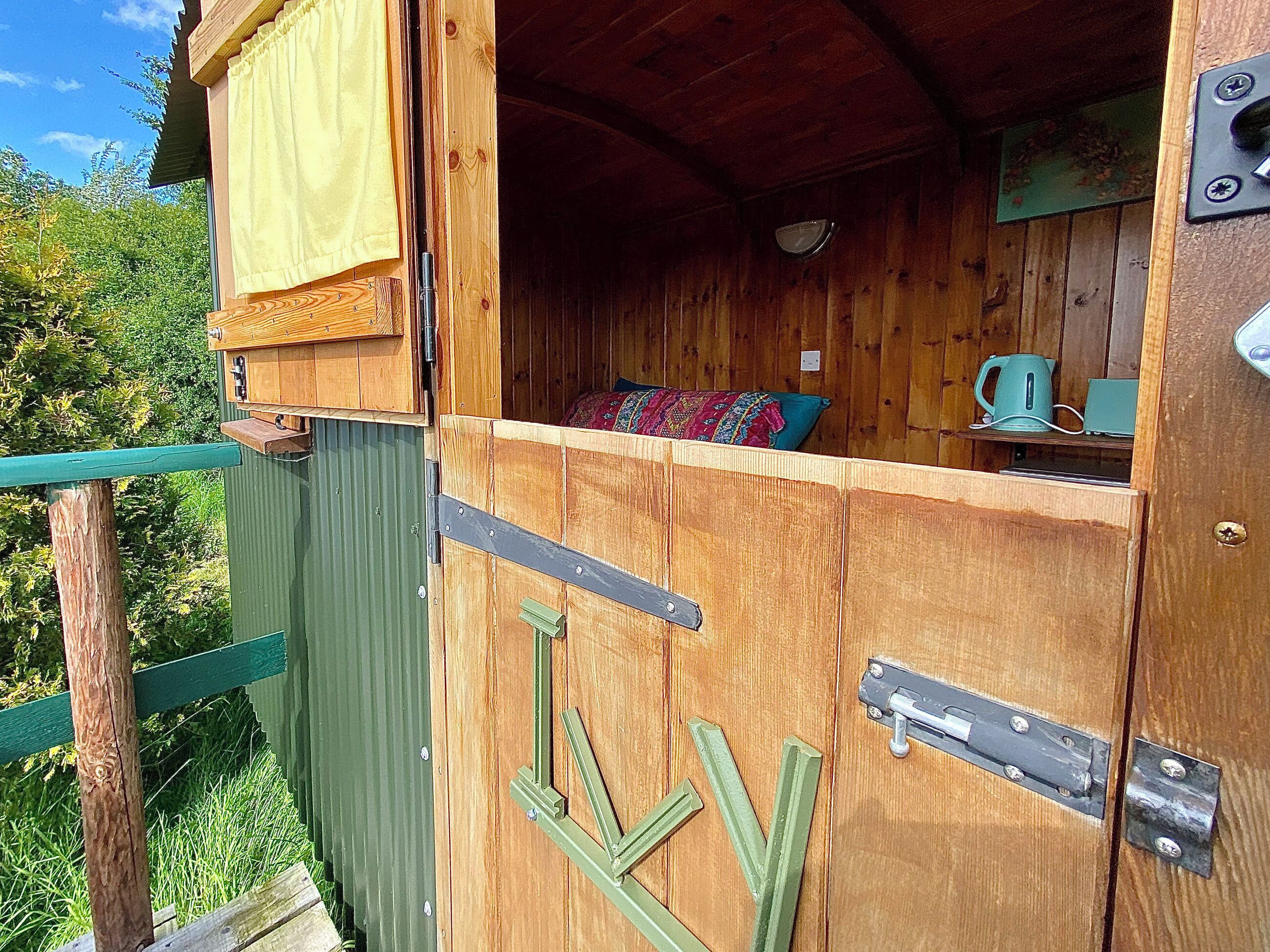 Eine Holzhütte mit grüner Fassade, ein sichtbares Bett mit bunter Decke und ein Wasserkocher auf einem Regal.