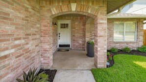 A brick house with a covered porch, a front door, and a small garden area. - Hot Tub | Game Room | Sleeps 10 & Pets (Cedar Park)