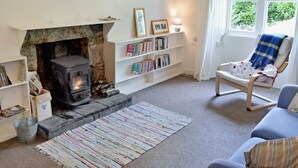 A cozy living room with a stone fireplace, a bookshelf, a comfortable chair, and a view of greenery outside. - A rural, detached cottage within the historic grounds of the Bargaly Estate. (Palnure near Newton Stewart)