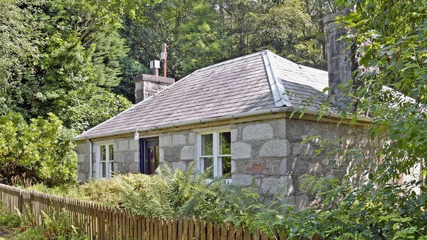 A stone house with a sloped roof, surrounded by dense greenery and a wooden fence. - A rural, detached cottage within the historic grounds of the Bargaly Estate. (Palnure near Newton Stewart)