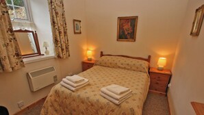 Un letto rifatto con una coperta a fantasia e due asciugamani piegati, una lampada da comodino, un quadro appeso al muro e una finestra con tende.