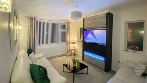 Una sala de estar moderna con un sofá blanco, una mesa de centro con una planta, un televisor montado en la pared y una ventana con persianas.