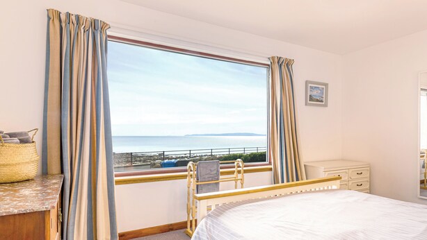A bedroom with a bed, a desk, a chair, a window with curtains, and a view of the sea.