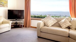 A living room with a sofa, a TV, and a large window with a view of the sea.