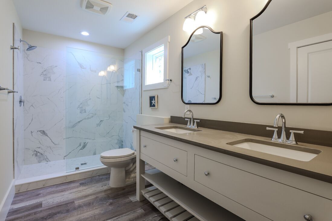 Un baño moderno con pared de ducha de mármol, un inodoro, un tocador con dos lavabos y un espejo grande.