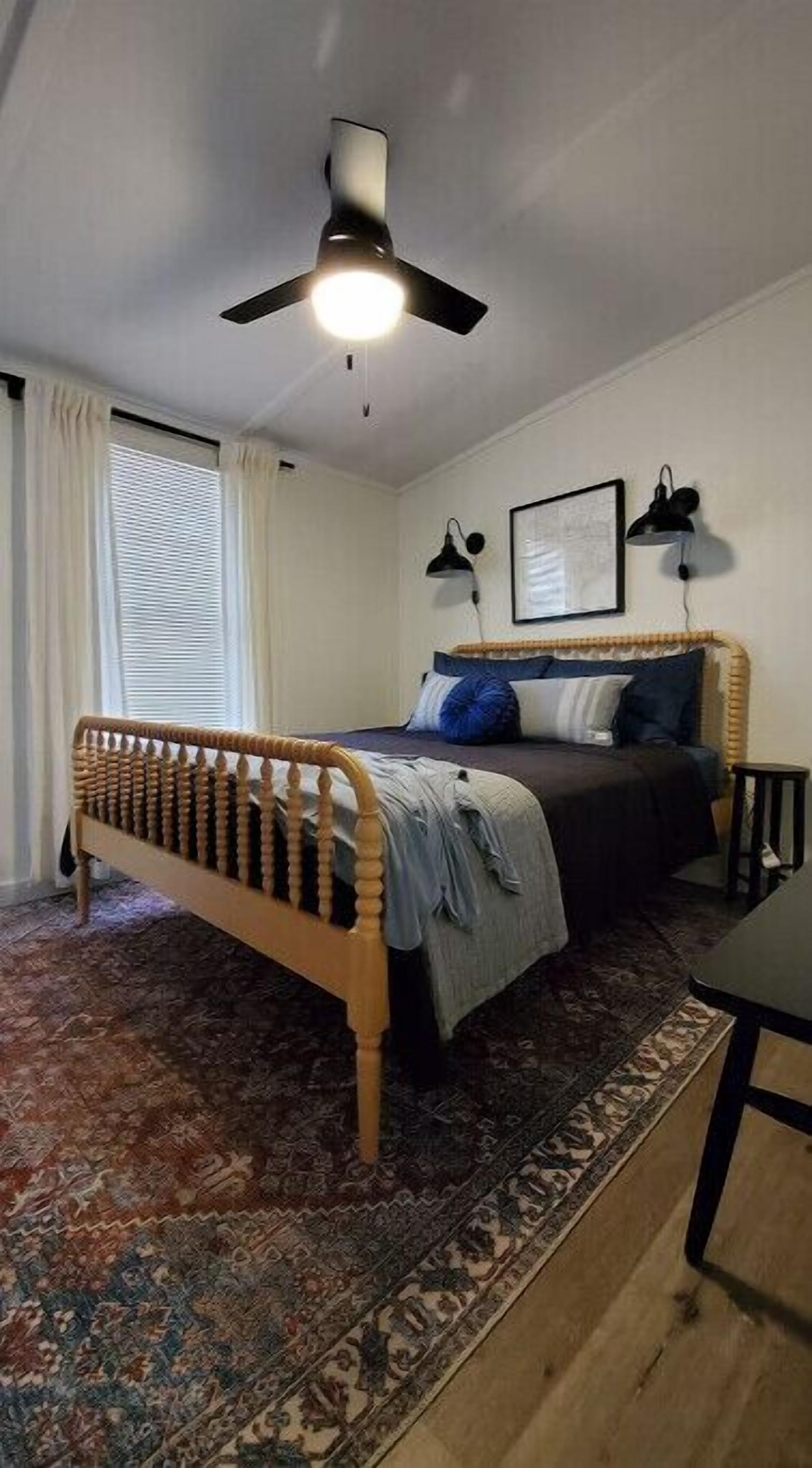 Une chambre à coucher comprenant un lit, un ventilateur de plafond, une fenêtre avec des rideaux, une lampe fixée au mur et un tapis.