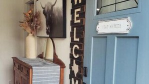 Een blauwe deur met een sterlogo, een 'WELCOME' bord, een klein krukje en een houten kastje met een gestreept kleedje.