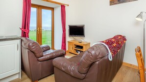 A room with a brown leather sofa, a television on a wooden stand, and a painting of a coastal landscape. - 2 bedroom accommodation in Mey, near Thurso (Mey, near Thurso)