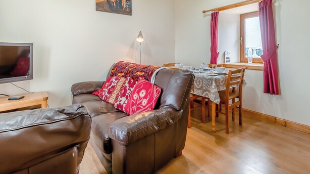 A cozy living room with a leather sofa, a dining table set for two, and a television on a wooden stand. - 2 bedroom accommodation in Mey, near Thurso (Mey, near Thurso)