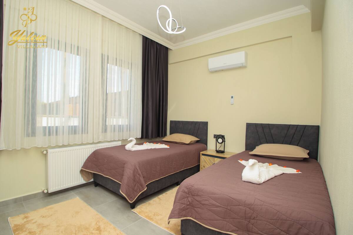 A hotel room with two beds, beige walls, a radiator, and a window with curtains.