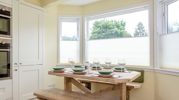 Una cocina con armarios blancos, una mesa de madera preparada para comer, y un ventanal con persianas.