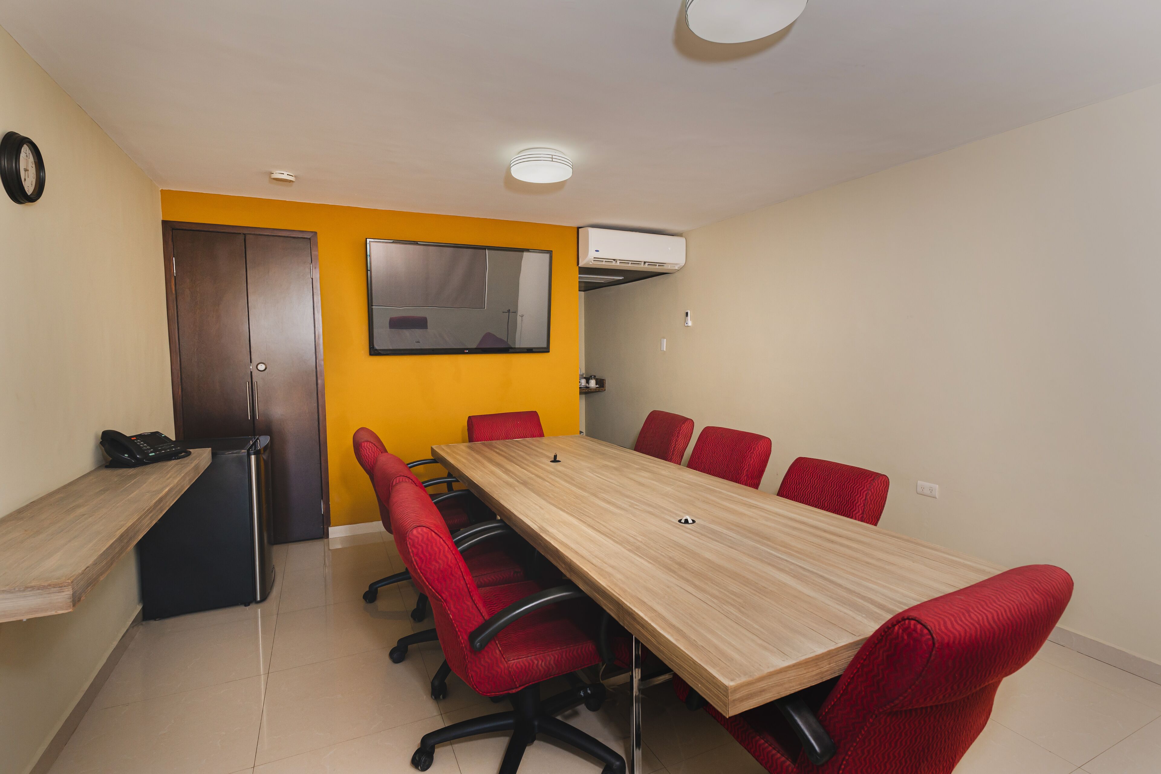 A conference room with a long table, red chairs, a wooden desk, a telephone, and an air conditioning unit.