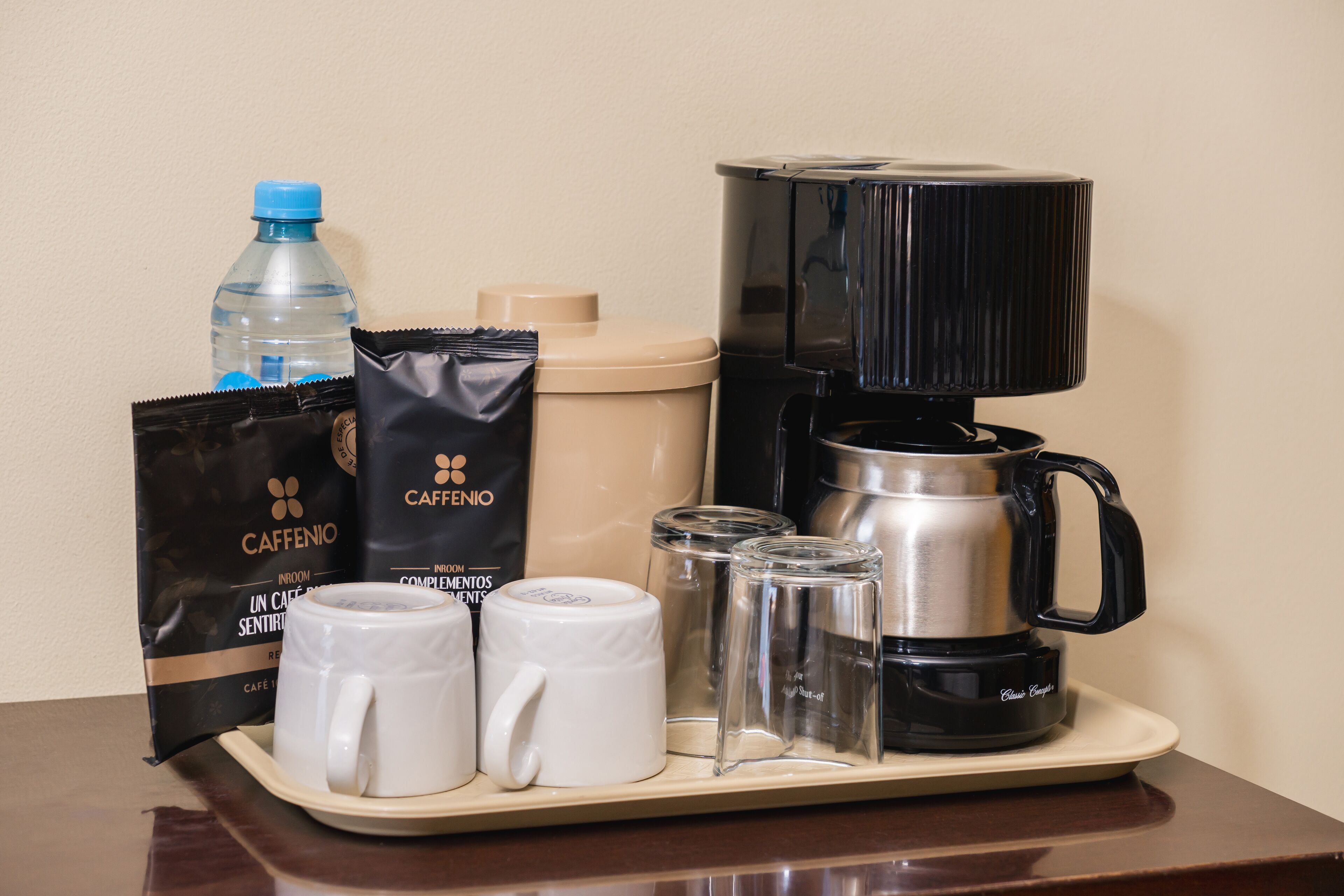 Un plateau contenant une machine à café, deux tasses blanches, une bouteille d’eau, deux paquets de café Caffenio et deux récipients en verre.