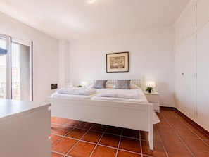 A bedroom with a white bed, a nightstand with a lamp, a framed picture on the wall, and a sliding door to a balcony.