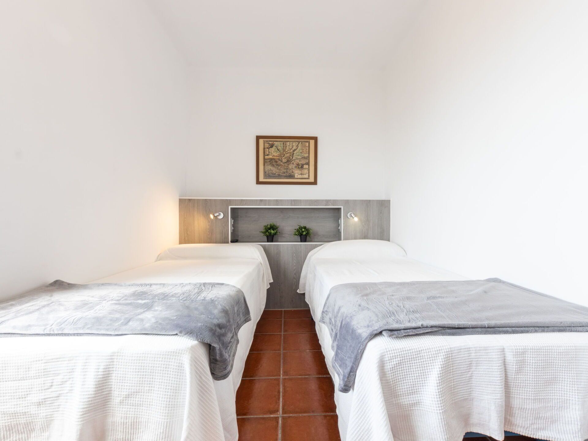 A room with two beds, a wooden headboard, a shelf with plants, and a framed picture on the wall.