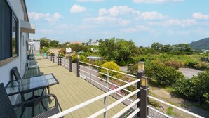 A balcony with a glass table and chairs, a wooden deck, and a view of a landscaped garden with trees and a hill in the distance. - Jeju Joyhouse (Seogwipo)