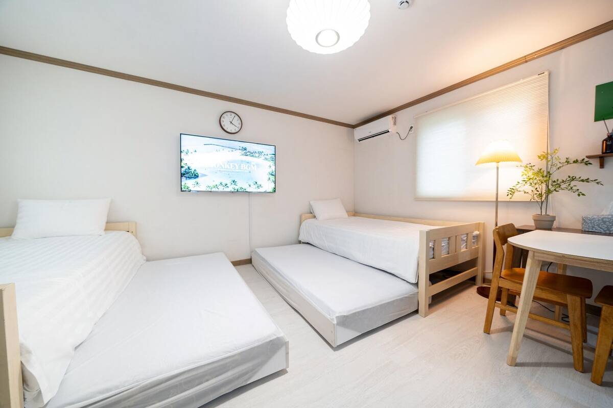 A room with three beds, a desk, a chair, a TV, and a window with blinds.