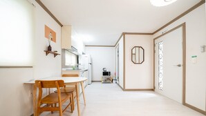 A modern kitchen with white appliances, a dining area with wooden chairs, and a hallway leading to a door with a decorative mirror. - Jeju Joyhouse (Seogwipo)