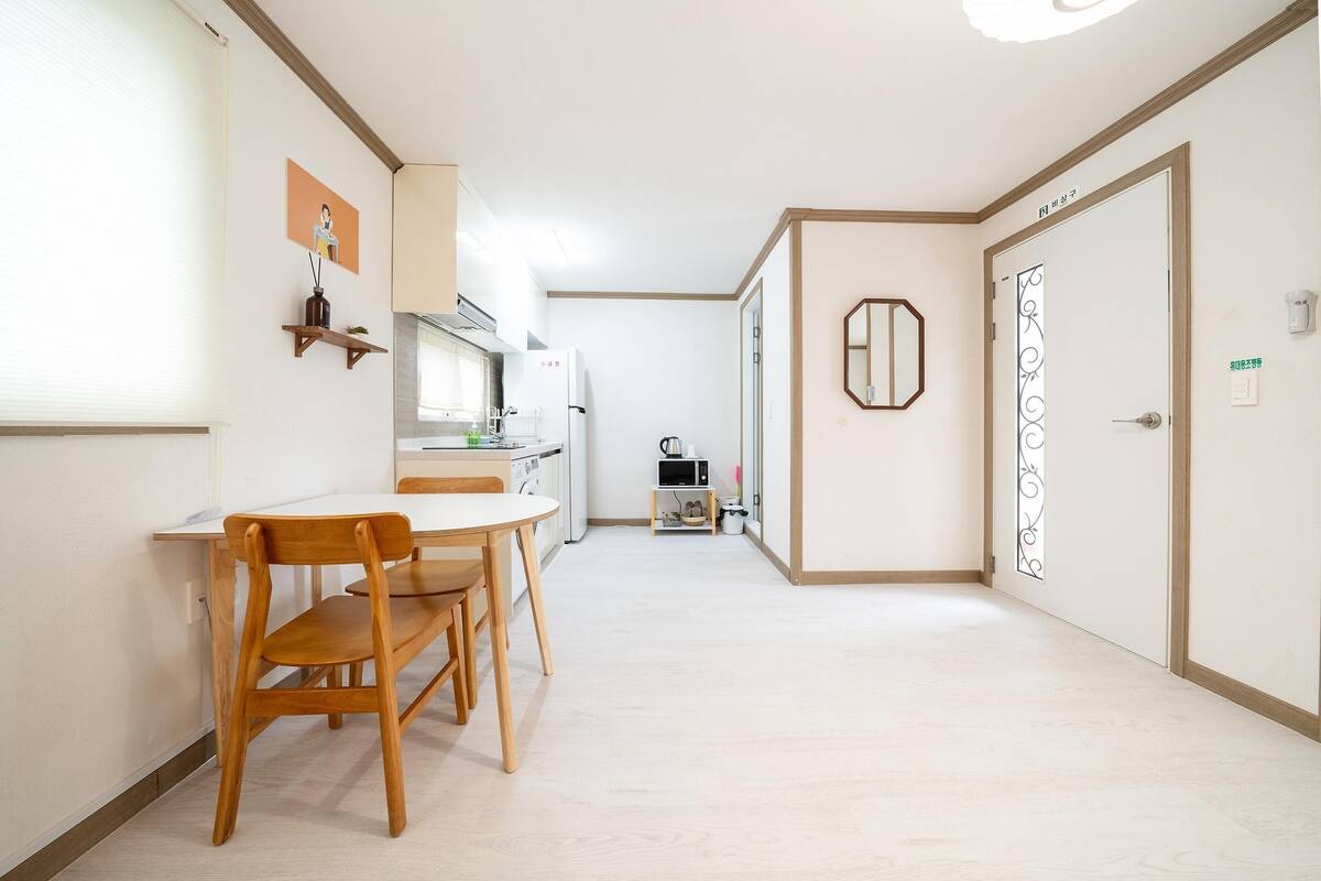 A modern kitchen with white appliances, a dining area with wooden chairs, and a hallway leading to a door with a decorative mirror.