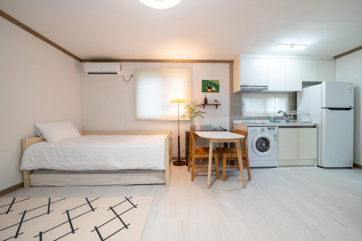 A compact room with a bed, a small table with chairs, a washing machine, and a refrigerator.