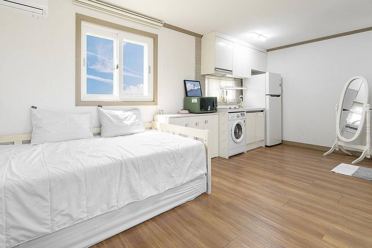 A bedroom with a bed, a washing machine, a refrigerator, and a bathroom mirror.