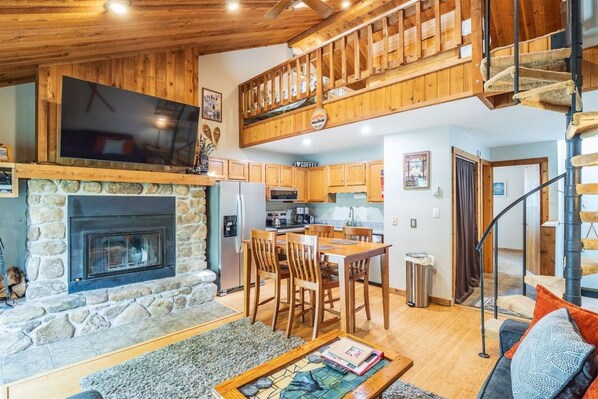 A cozy interior with a stone fireplace, wooden dining area, and a staircase leading to an upper level.